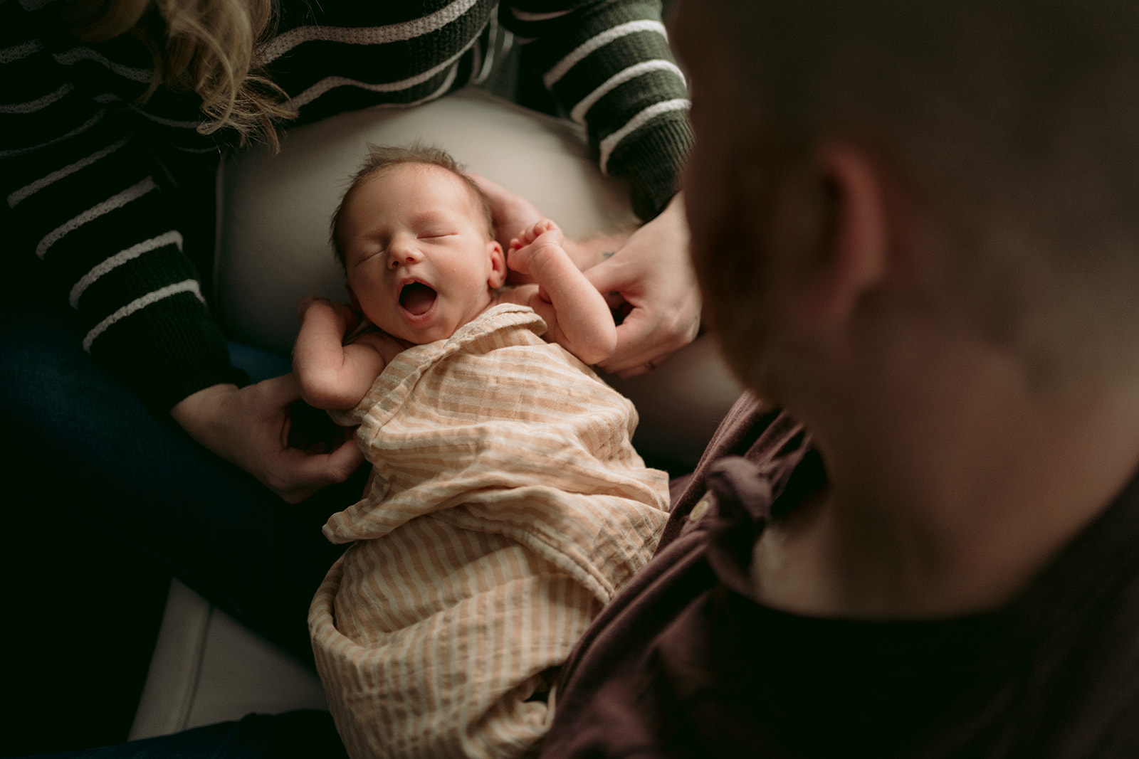 Newborn baby yawns in the window light being held by mother and father.