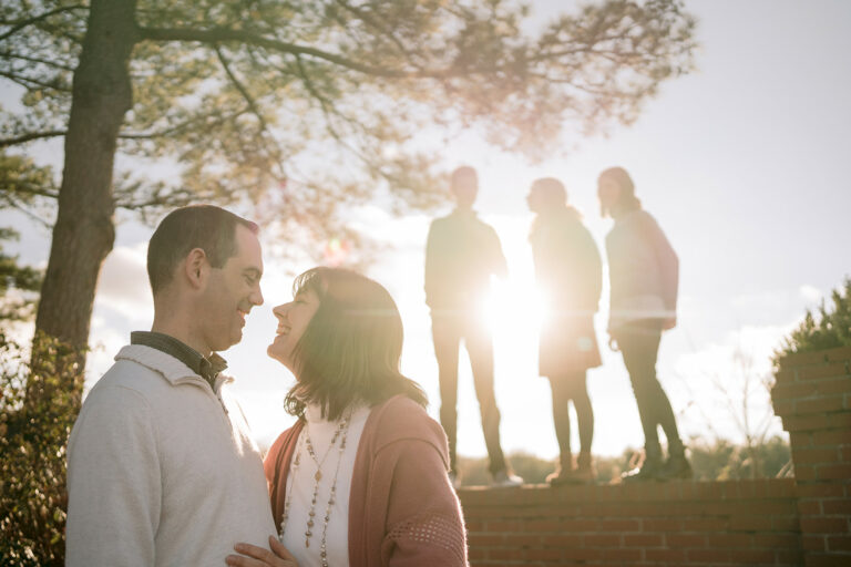Parents smile at each other as siblings stand behind them during an evening family photoshoot.