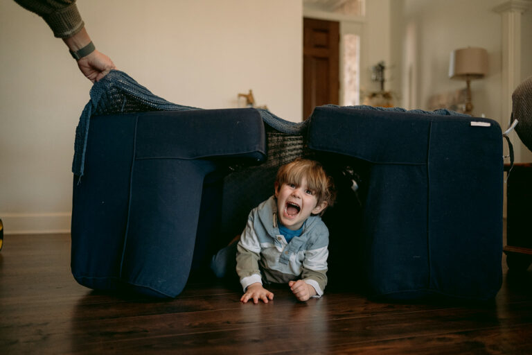 Little boy pokes out of a fort he made at home in the living room.