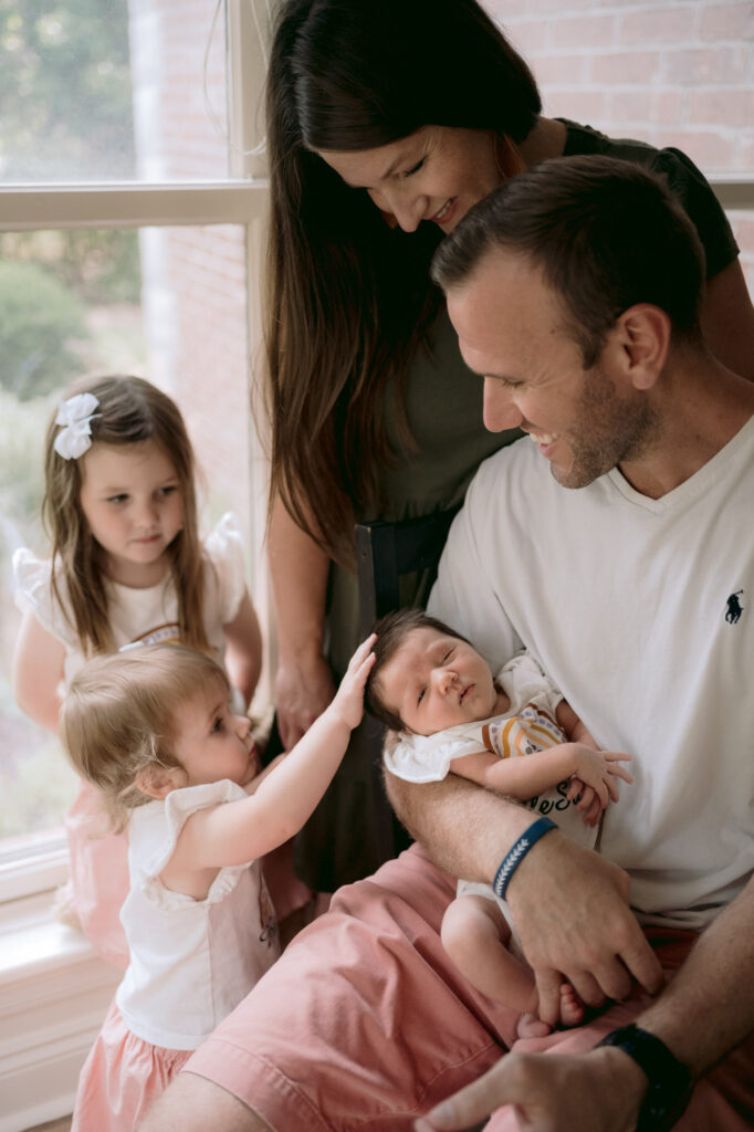 family sitting by a window looking at their newborn baby girl as sister touches her head